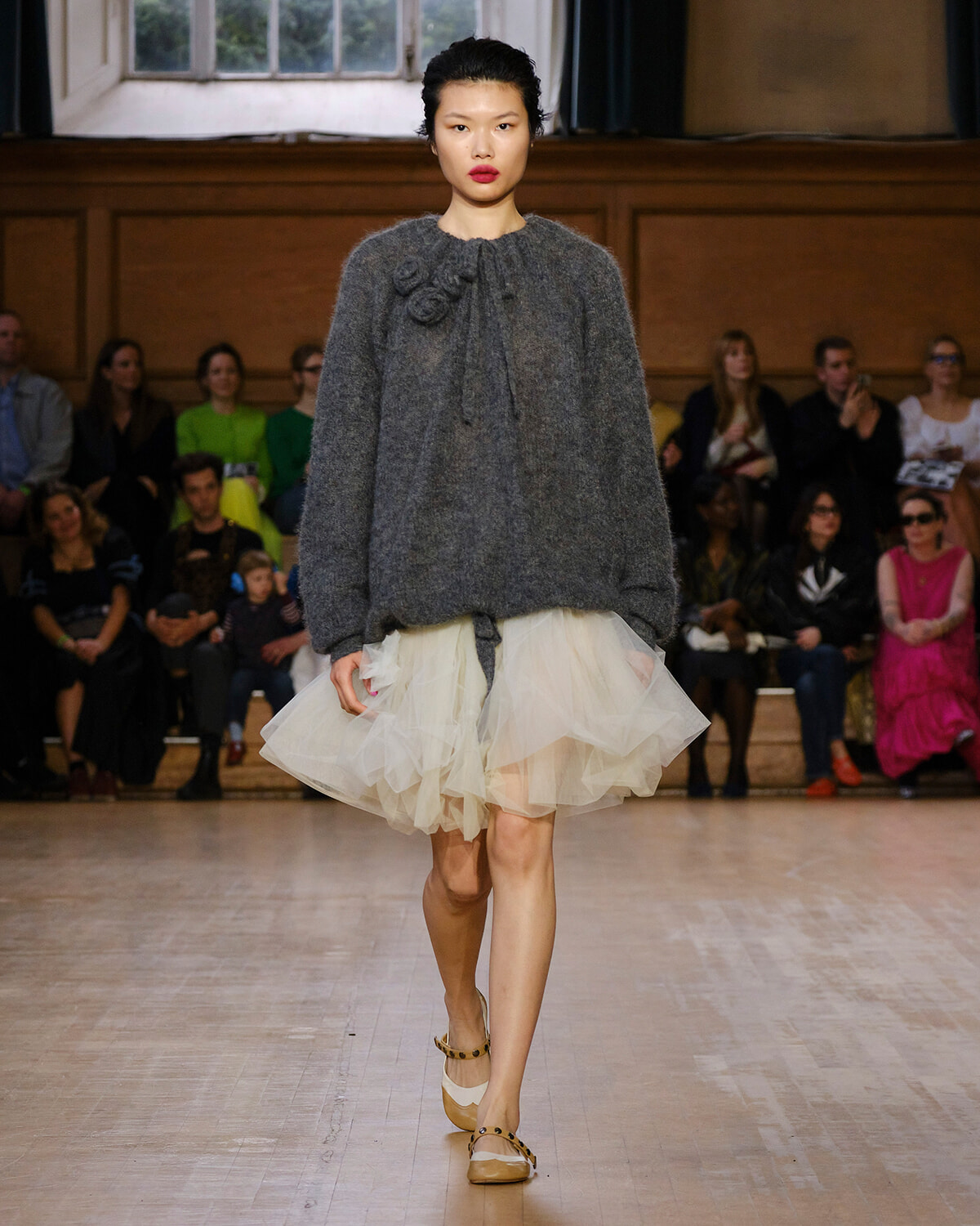 Model walking a fashion show in a chunky gray knit sweater with flower embellishments and a cream tulle skirt; audience in background.