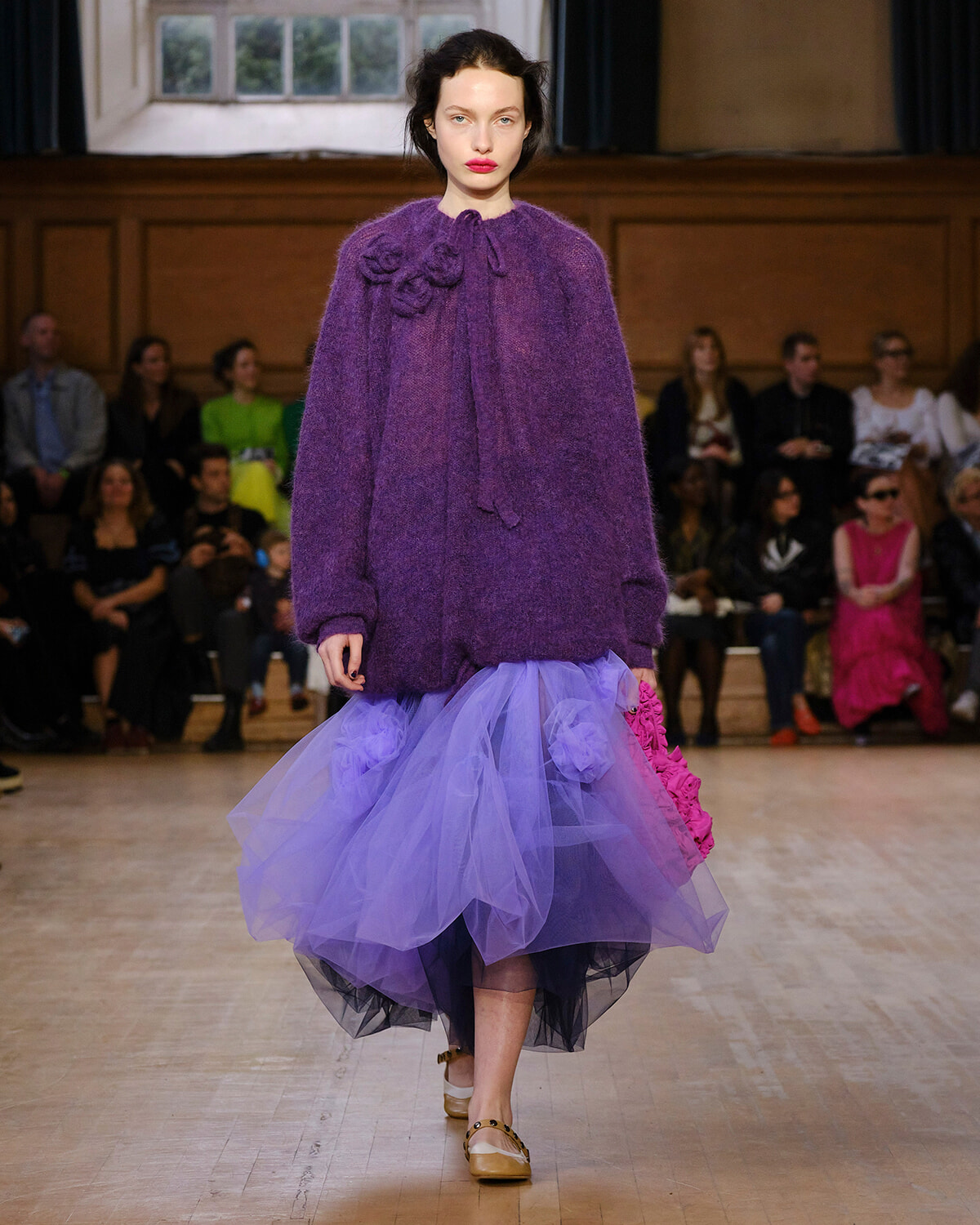 Model walking a runway in a chunky purple sweater and lavender tulle skirt, with a pink accessory in hand and an audience in the background.