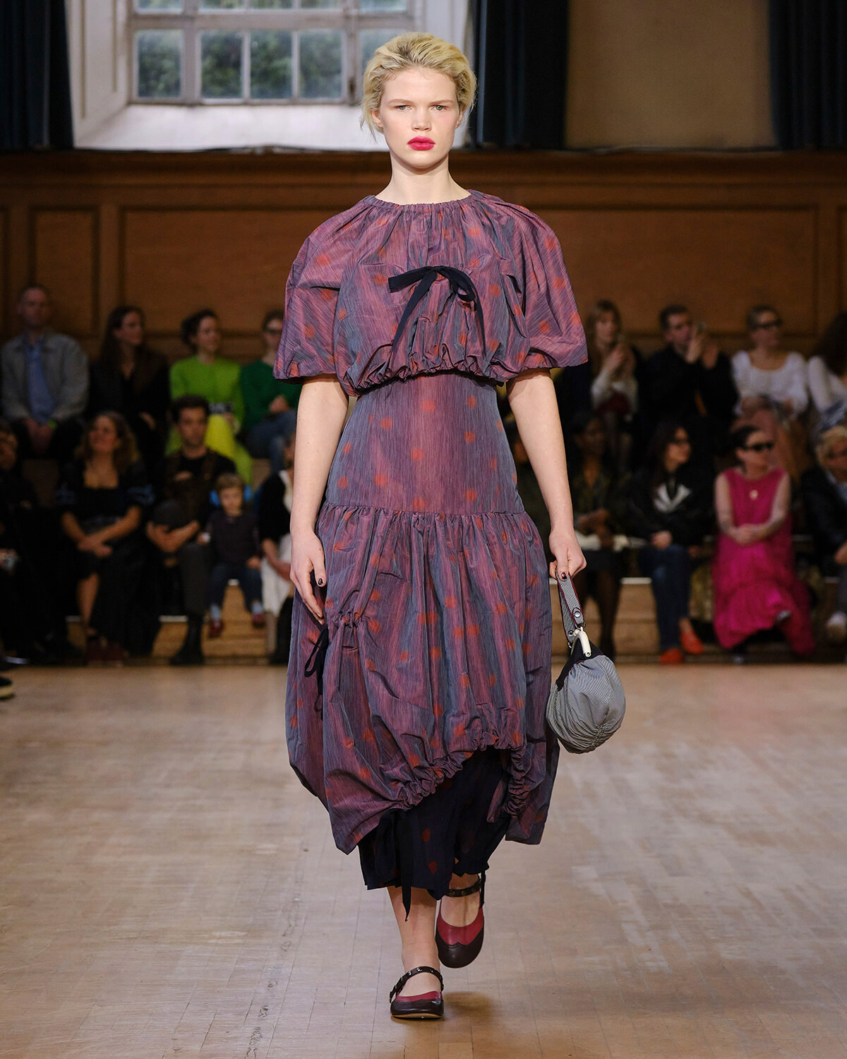 Model walking a fashion show in a purple dotted puff-sleeve dress with a black ribbon belt, carrying a gray handbag.