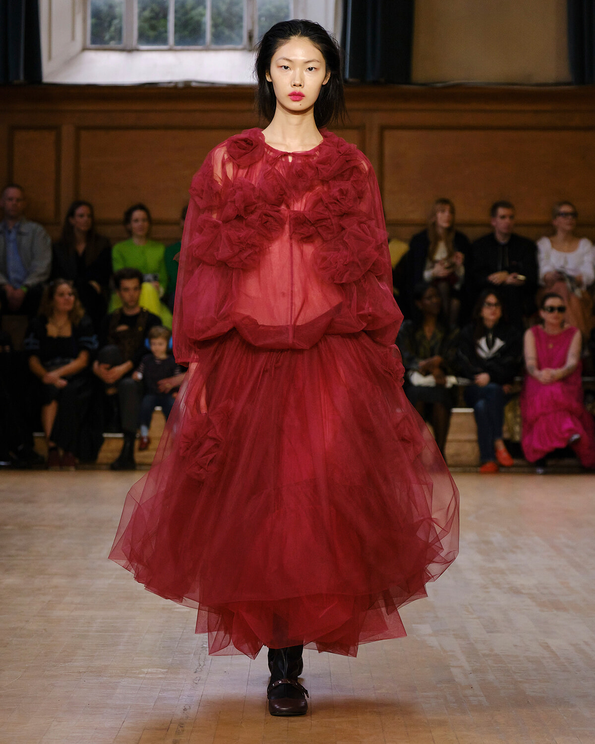 Model walks the runway in a voluminous red tulle gown with layered ruffles and sheer overlay, audience watching in the background.