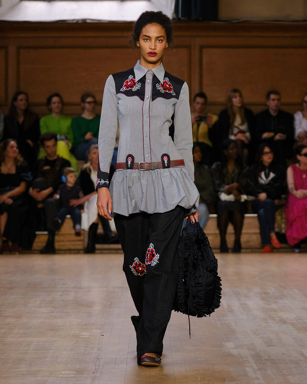 Runway model in a gray striped button-up shirt with red floral embroidery on the yoke and cuffs, paired with dark jeans with floral patches, walking the catwalk.