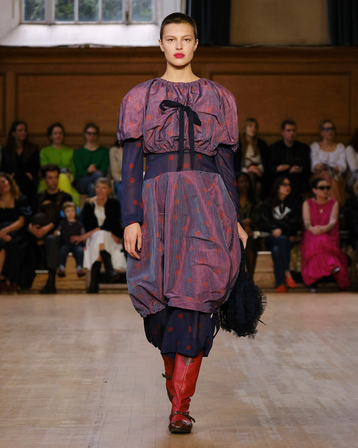 Model walks the runway in a voluminous purple-pink polka-dot dress with a black bow, paired with red knee-high boots, audience in the background.] ,need to fix