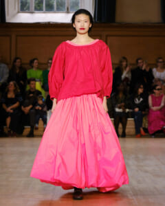Model on runway wearing a bright pink off-shoulder blouse and voluminous skirt, audience in background watching fashion show