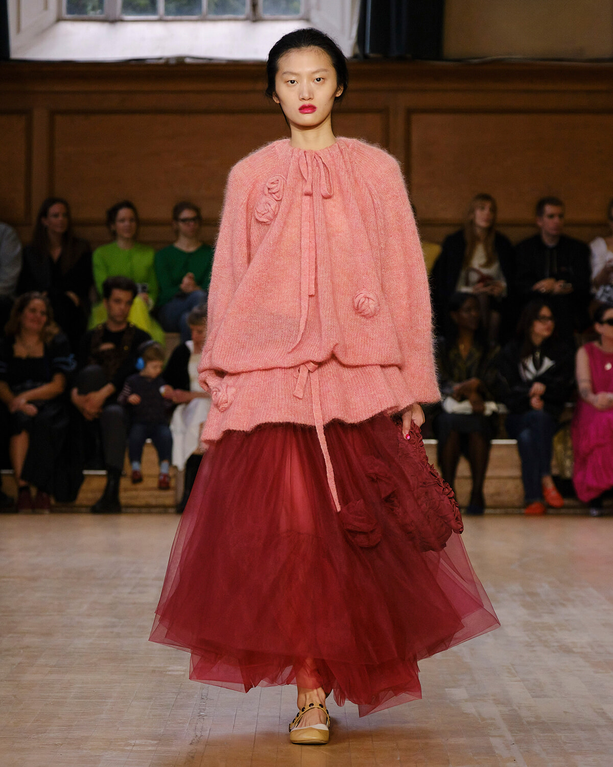 Runway model wearing a pink fuzzy cape with gathered bow ties and a burgundy tulle skirt; audience watches in the background.