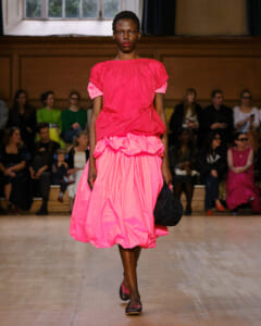 Model walking a runway in a bright pink, voluminous dress with ruffled hem, holding a black bag; audience seated in background as spectators watch.