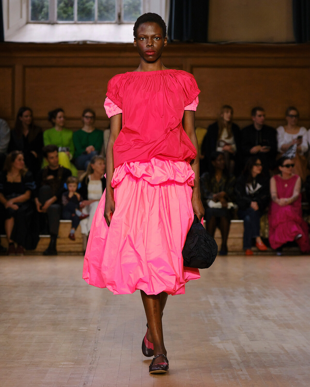 Model walking a runway in a bright pink, voluminous dress with ruffled hem, holding a black bag; audience seated in background as spectators watch.
