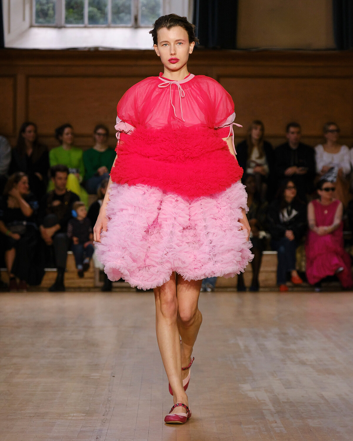 Model walks down runway in a bright pink sheer top with a magenta fluffy pink layered dress; audience seated along the sides.
