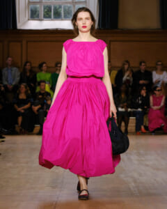 Model walks a runway in a bright pink, full-skirt dress; audience sits along the sides watching the show in a large room.