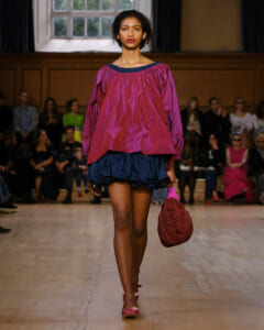Model walks a runway in a bright magenta off-shoulder top with a navy blue ruffled skirt, carrying a maroon handbag and wearing pink heels.
