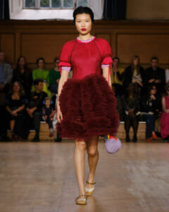 Runway model in a red sheer blouse and voluminous burgundy ruffled skirt graces the fashion show catwalk with an audience in the background.