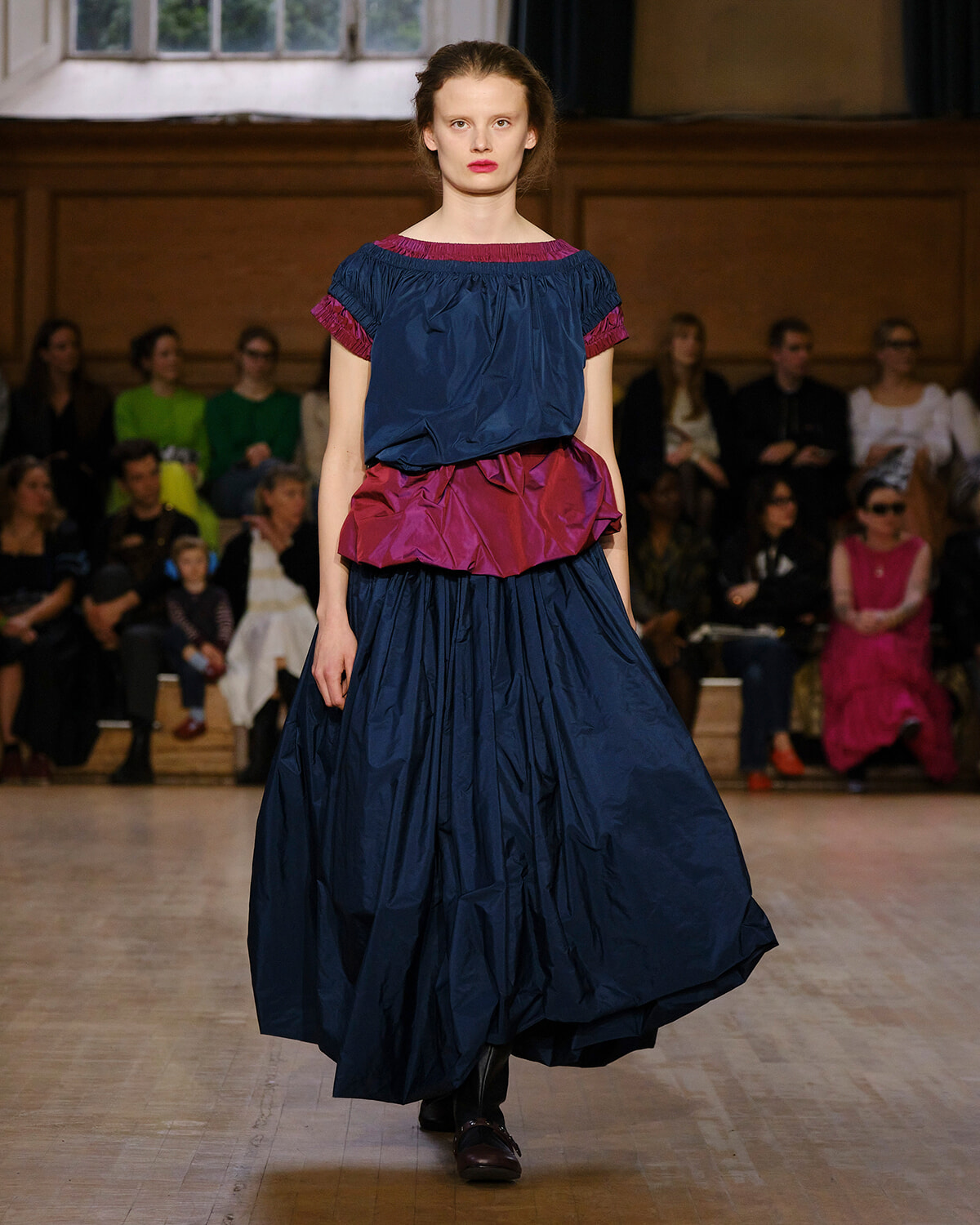 Model walks a fashion runway in a navy blue gown with a magenta ruffle waist, audience seated along the backdrop.