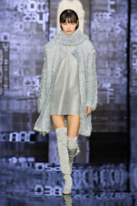 Model on runway wearing a textured silver coat over a light gray dress with silver knee-high boots.
