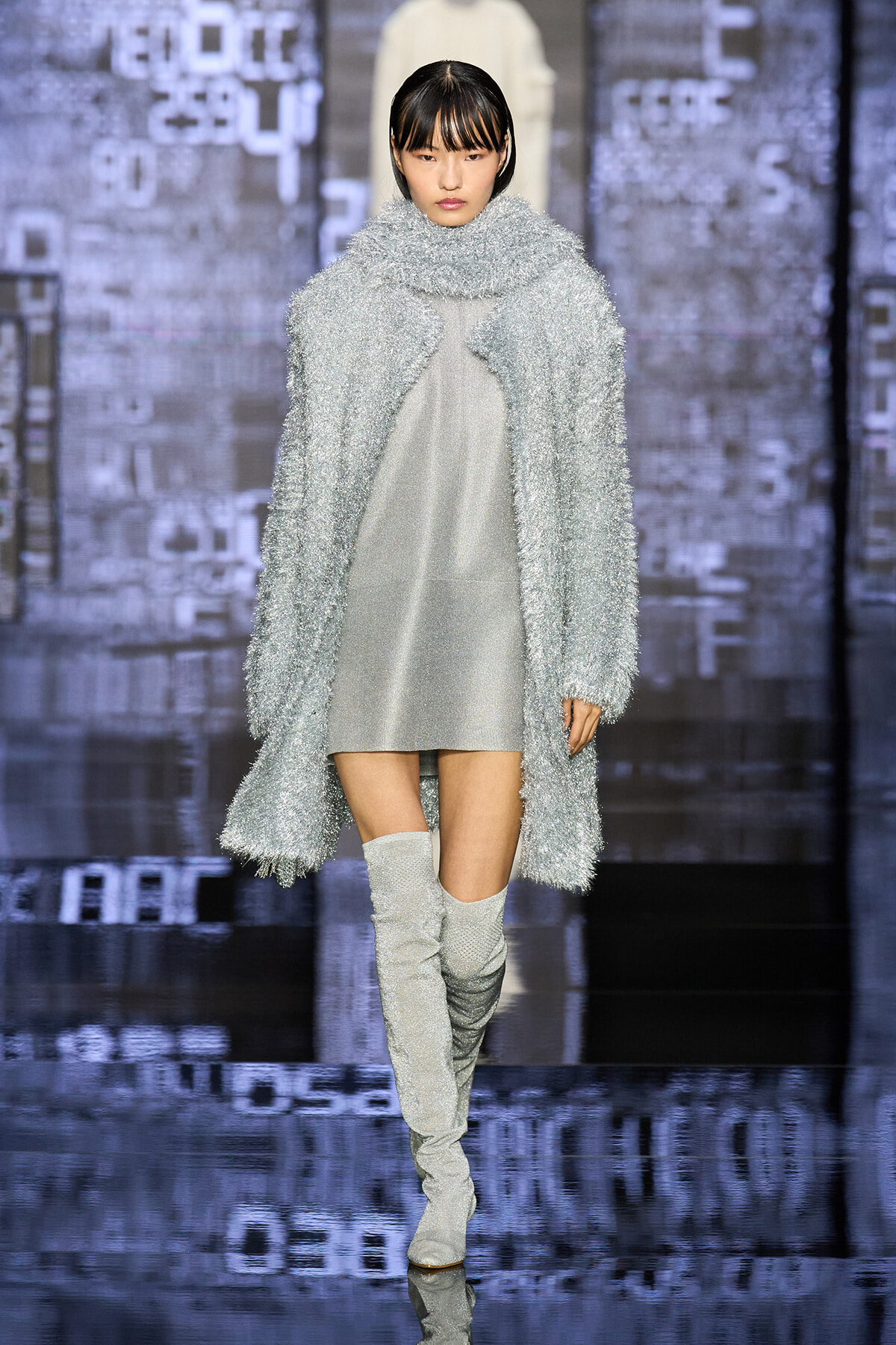 Model on runway wearing a textured silver coat over a light gray dress with silver knee-high boots.