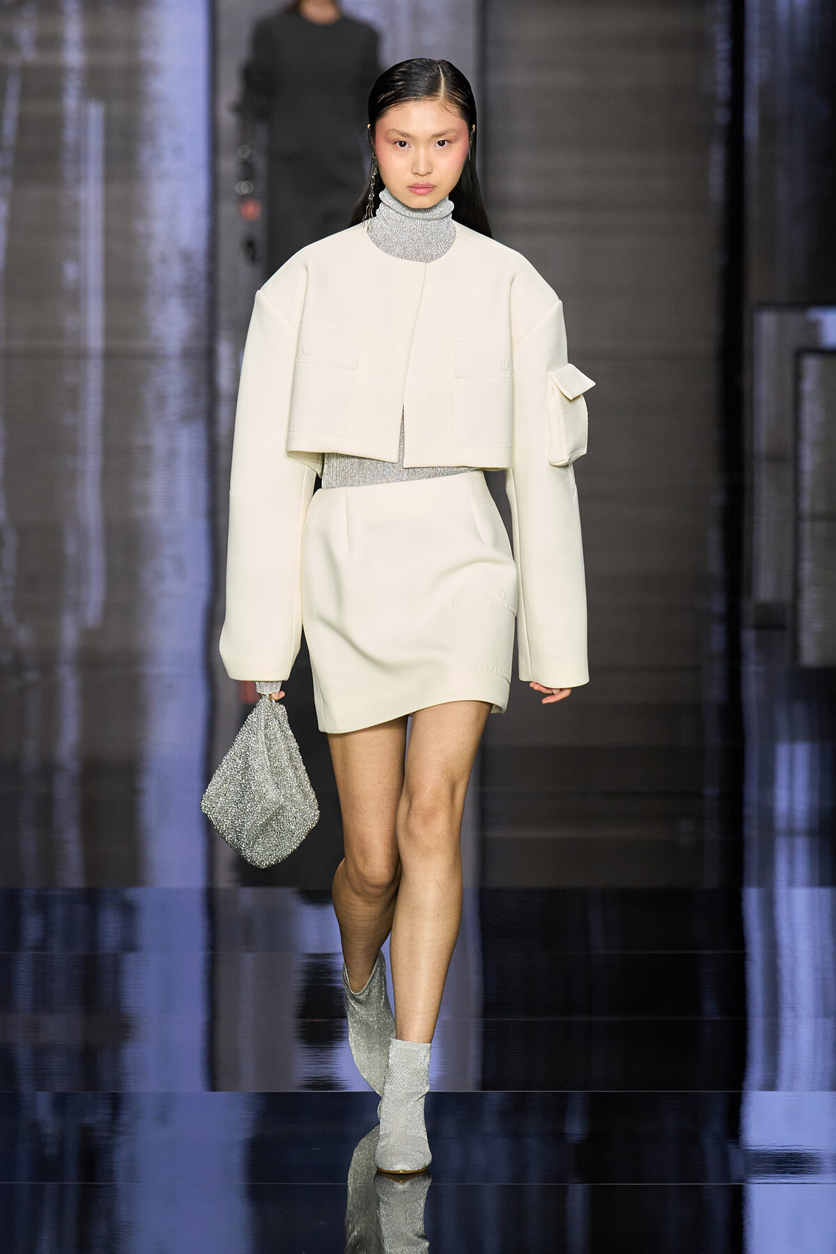 Model walking the runway in an all-white ensemble: cropped jacket over a metallic turtleneck and matching mini skirt, with silver glitter boots and a small glitter clutch.