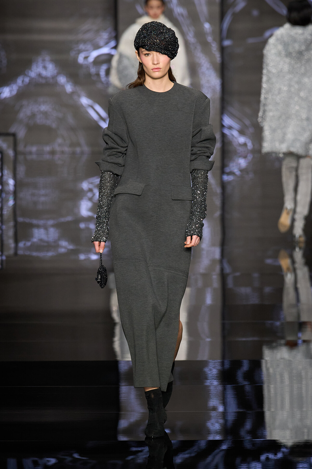 Model walking a runway in a dark gray, structured knit suit with puffed sleeves, glittery long gloves, and a glittery cap highlighted against a reflective backdrop.