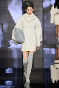 Model on a runway wearing an oversized white knit sweater with a chunky scarf, holding a silver metallic bag, and glittery thigh-high boots.
