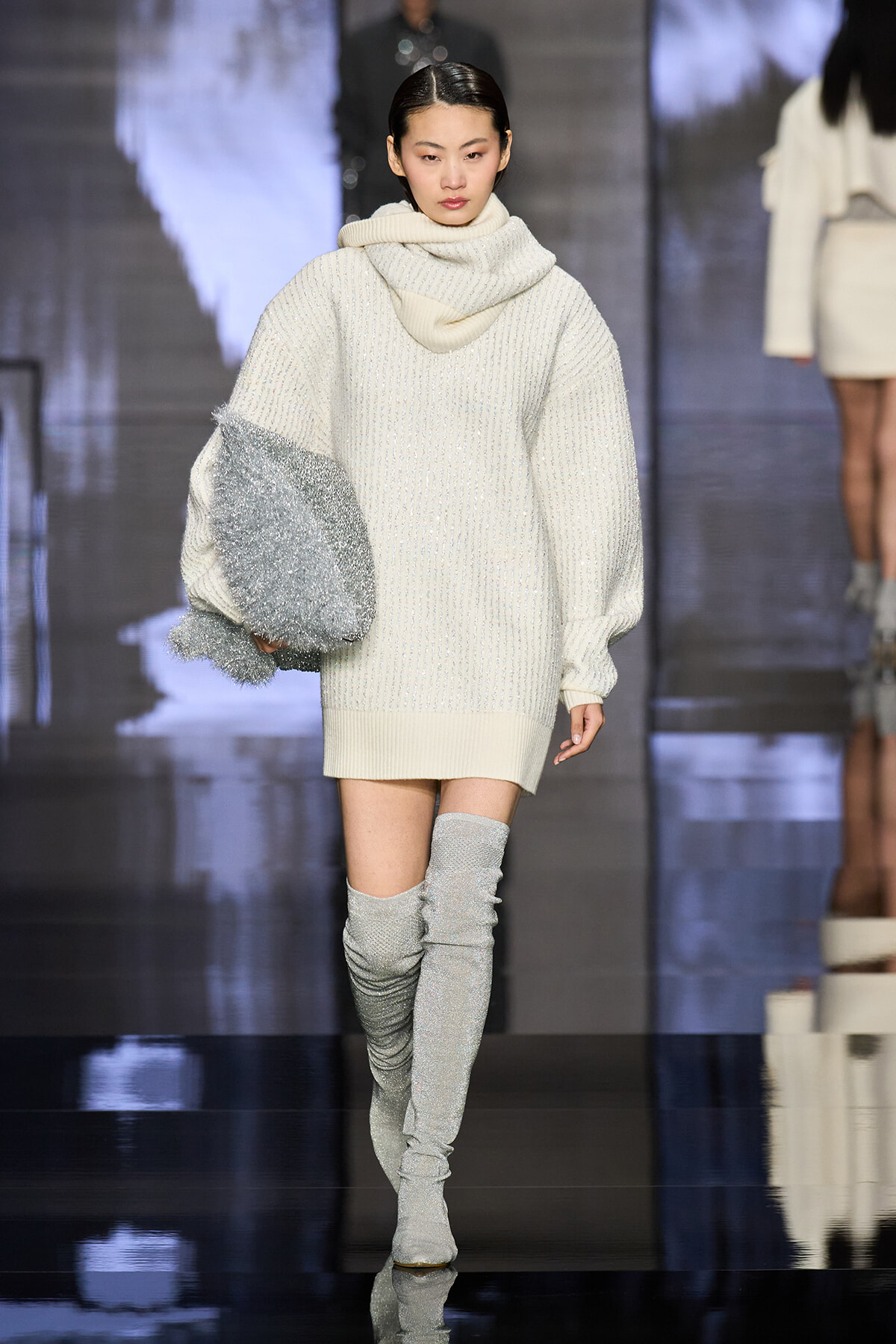 Model on a runway wearing an oversized white knit sweater with a chunky scarf, holding a silver metallic bag, and glittery thigh-high boots.