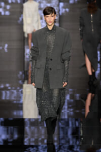 Model on a runway wearing a loose gray blazer over a sparkling silver dress, with black boots and tights, walking the catwalk.
