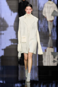 Model walks runway in ivory knit coat with wide lapels and matching turtleneck, small front pocket, and sparkly silver ankle boots.