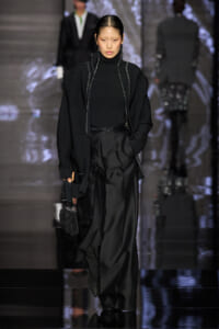 Model walks the runway in an all-black ensemble: oversized knit cardigan over a black turtleneck and wide-leg satin pants, carrying a small beaded black bag.
