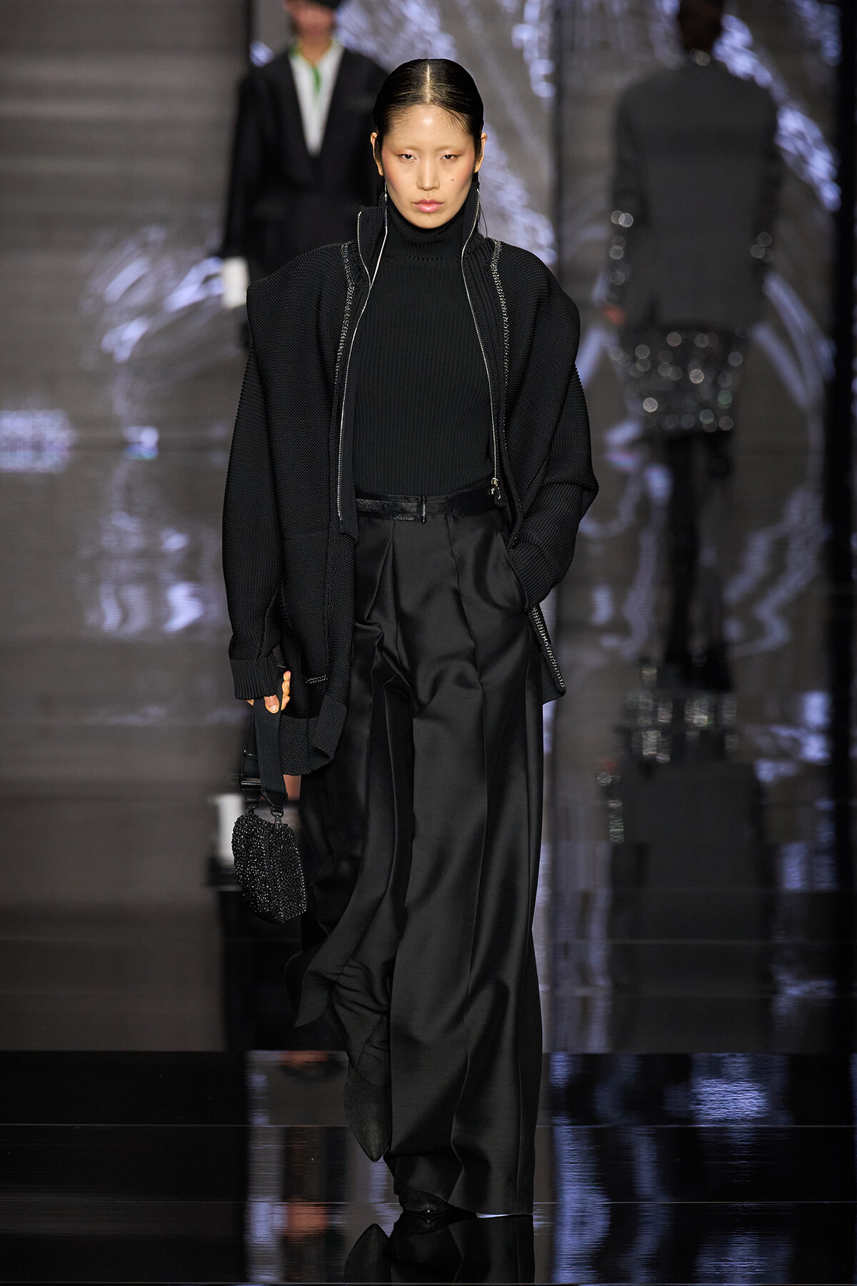 Model walks the runway in an all-black ensemble: oversized knit cardigan over a black turtleneck and wide-leg satin pants, carrying a small beaded black bag.