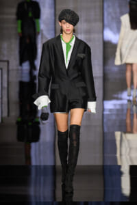Model on a fashion runway wearing a black blazer with green inner top, white shirt cuffs, a black sparkly beret, and thigh-high boots, holding a small beaded bag.