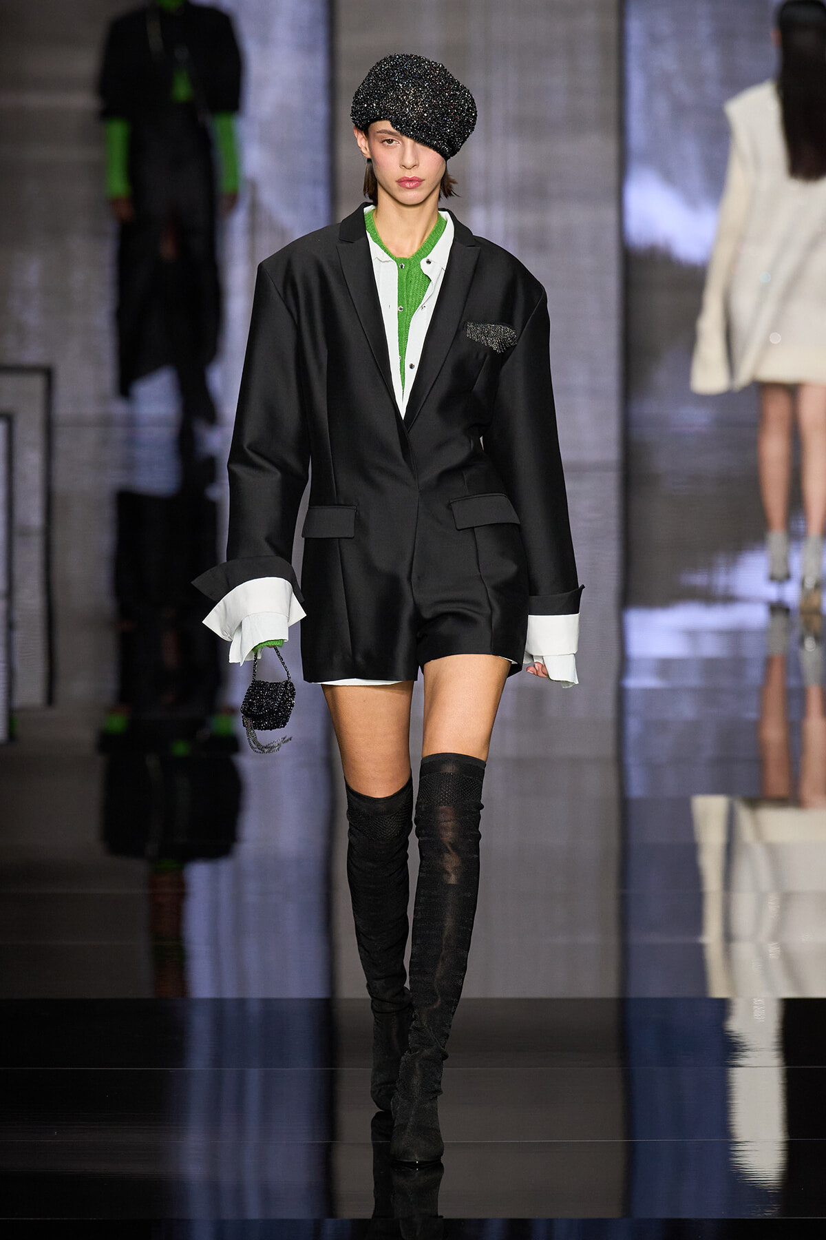 Model on a fashion runway wearing a black blazer with green inner top, white shirt cuffs, a black sparkly beret, and thigh-high boots, holding a small beaded bag.