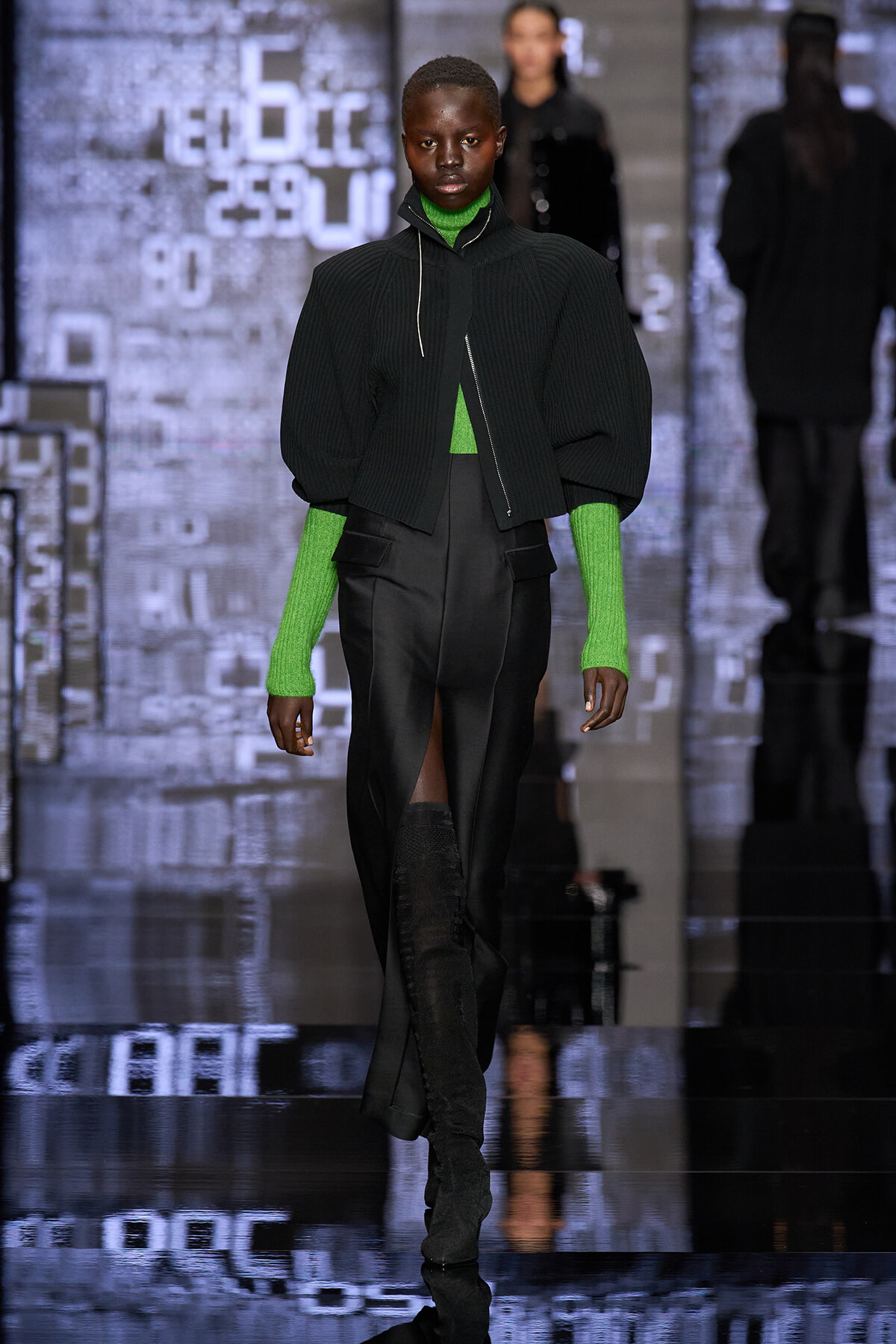 Runway model wearing a black ribbed cropped jacket over a bright green turtleneck and matching green sleeves with black high-waisted pants and a front slit.