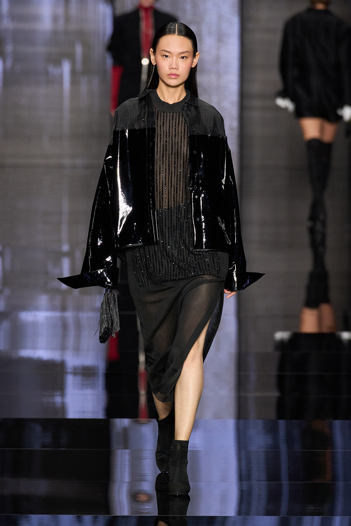 Runway model wearing a glossy black patent jacket over a sheer black beaded top and a black skirt, holding a small tassel bag on a reflective catwalk.