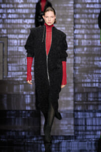 Model walking a runway in a glittery black zip-front suit with red turtleneck sleeves