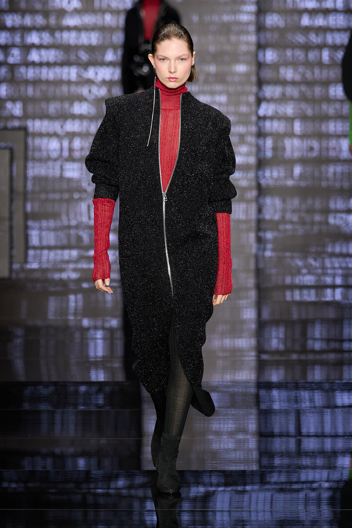 Model walking a runway in a glittery black zip-front suit with red turtleneck sleeves