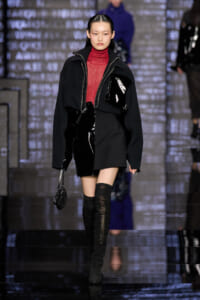 Model on a runway wearing a black oversized jacket with a red turtleneck and black knee-high boots, against a metallic backdrop.