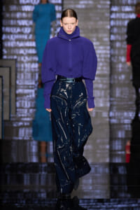 Model on a runway wearing a purple knit turtleneck with oversized sleeves and glossy navy pants on a fashion catwalk.