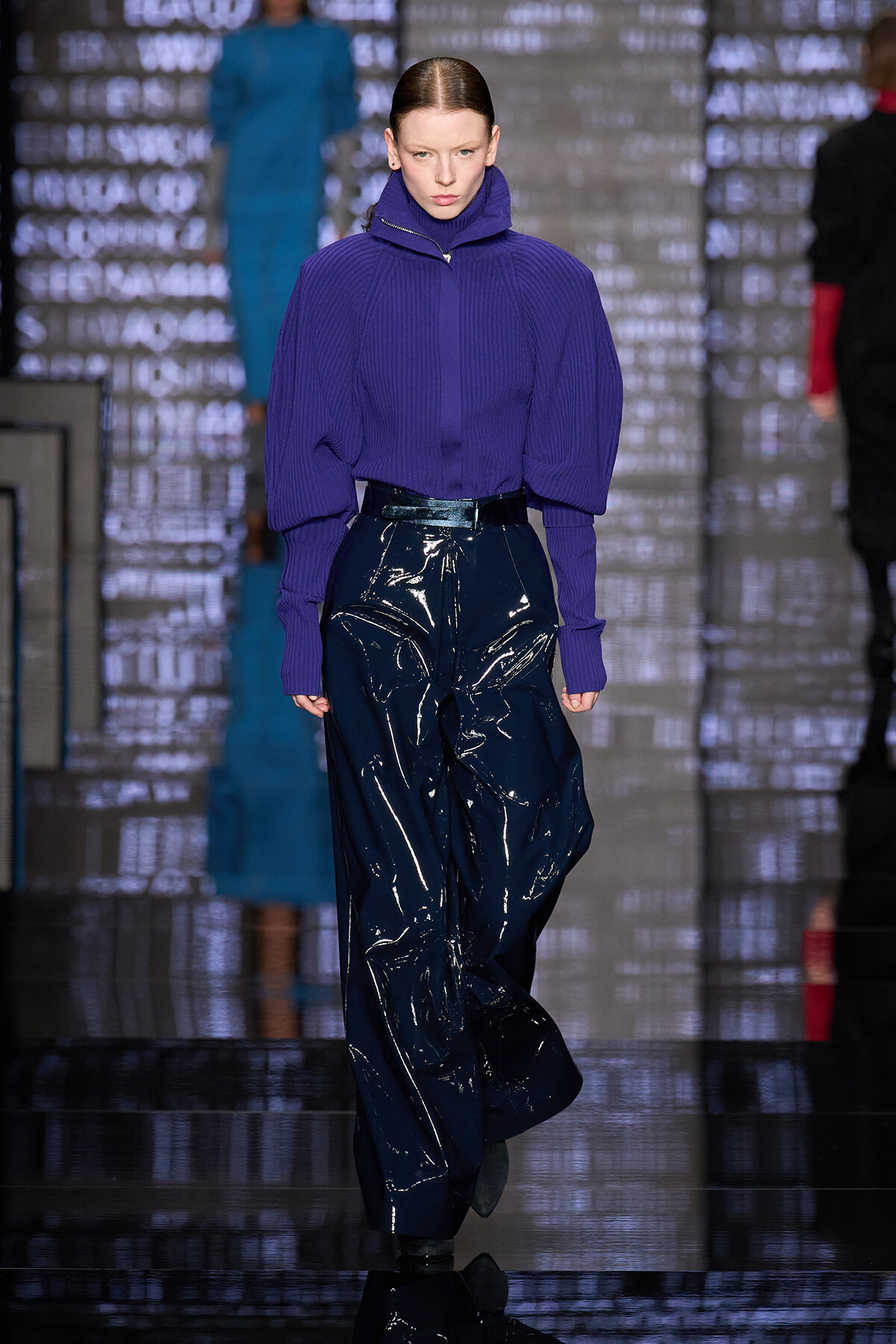 Model on a runway wearing a purple knit turtleneck with oversized sleeves and glossy navy pants on a fashion catwalk.