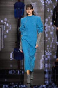 Model walking a runway in a turquoise glittery outfit with oversized sleeves and silver glitter gloves, paired with gray ankle boots.