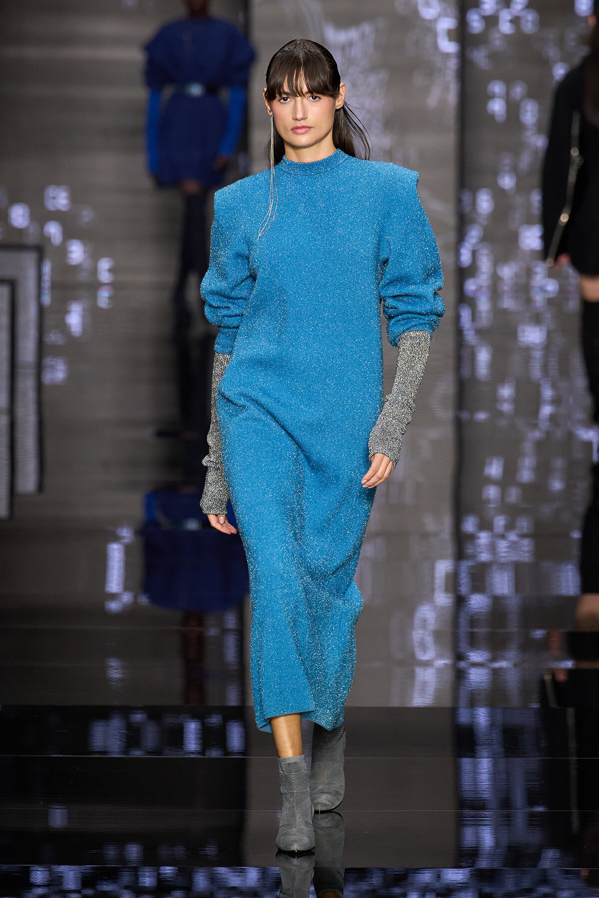 Model walking a runway in a turquoise glittery outfit with oversized sleeves and silver glitter gloves, paired with gray ankle boots.