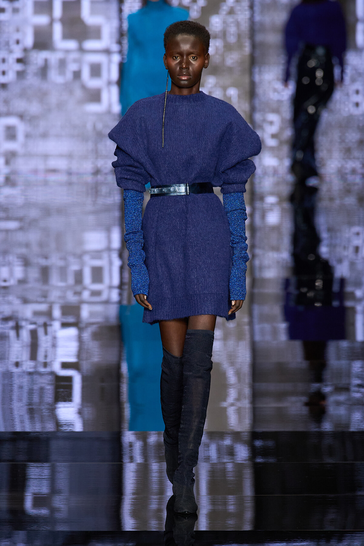 .Model walking a runway in a cobalt-blue knit sweater dress with a shiny black belt and sparkly blue gloves, tall black boots, blurred digital backdrop behind.