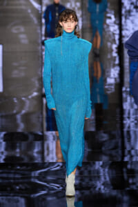 Model walks a glittery blue turtleneck gown on a reflective runway; silver boots complete the look.