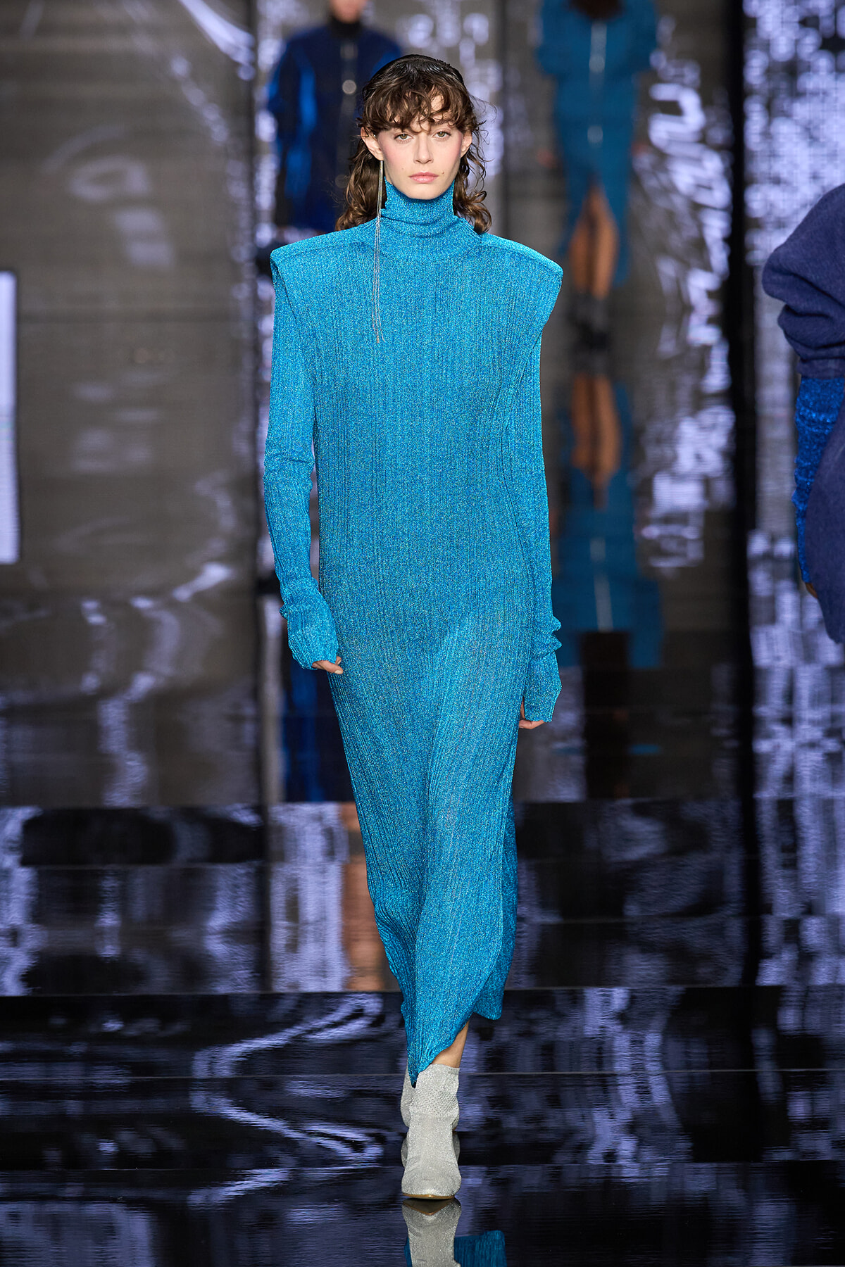 Model walks a glittery blue turtleneck gown on a reflective runway; silver boots complete the look.