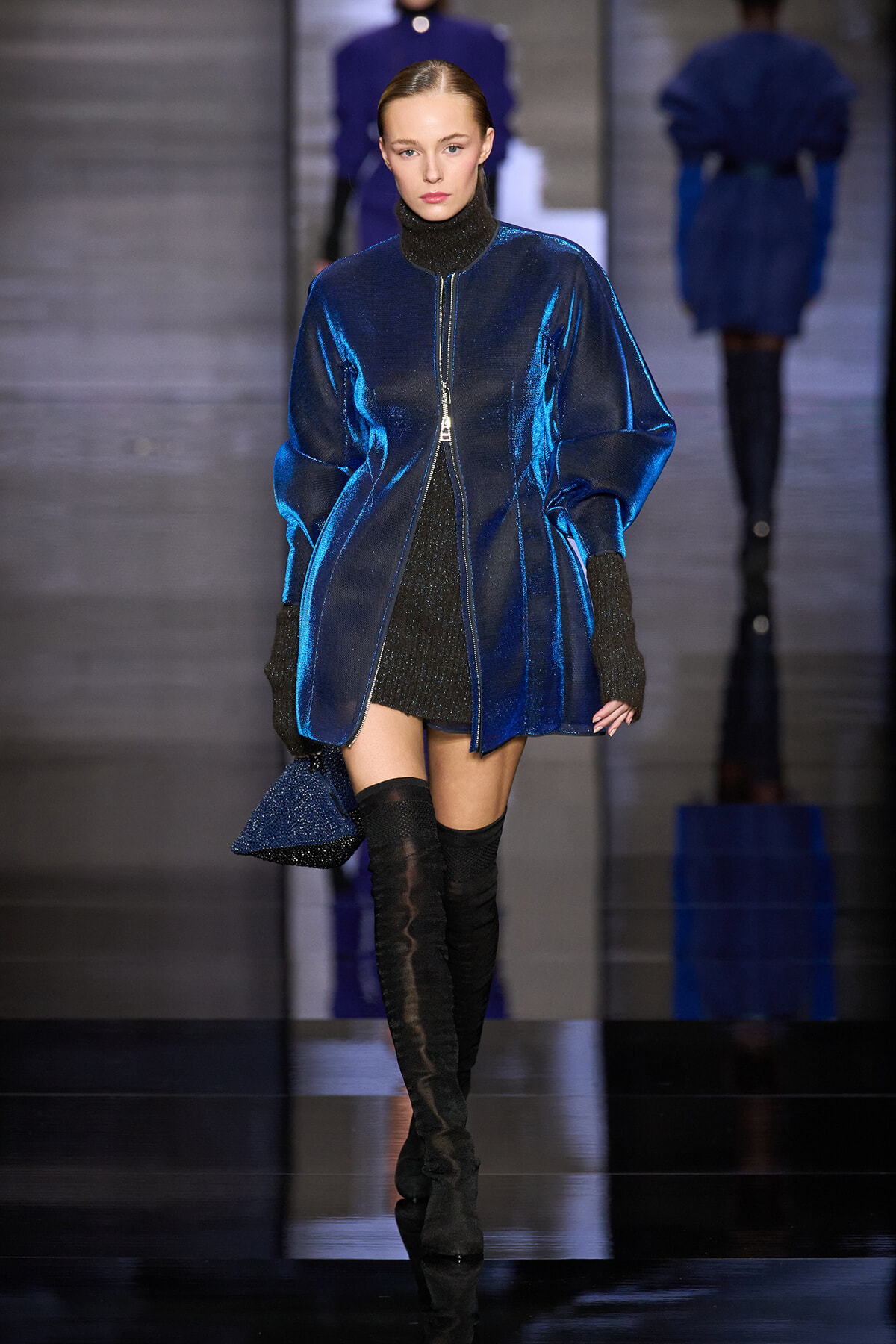 Model on a fashion runway wearing a metallic cobalt blue zip-front coat with oversized sleeves over a black turtleneck and glittery dress, paired with black thigh-high boots and gloves.