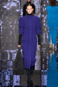 Model walking runway in a vivid purple knit midi dress with black turtleneck sleeves, holding a sparkly black bag.
