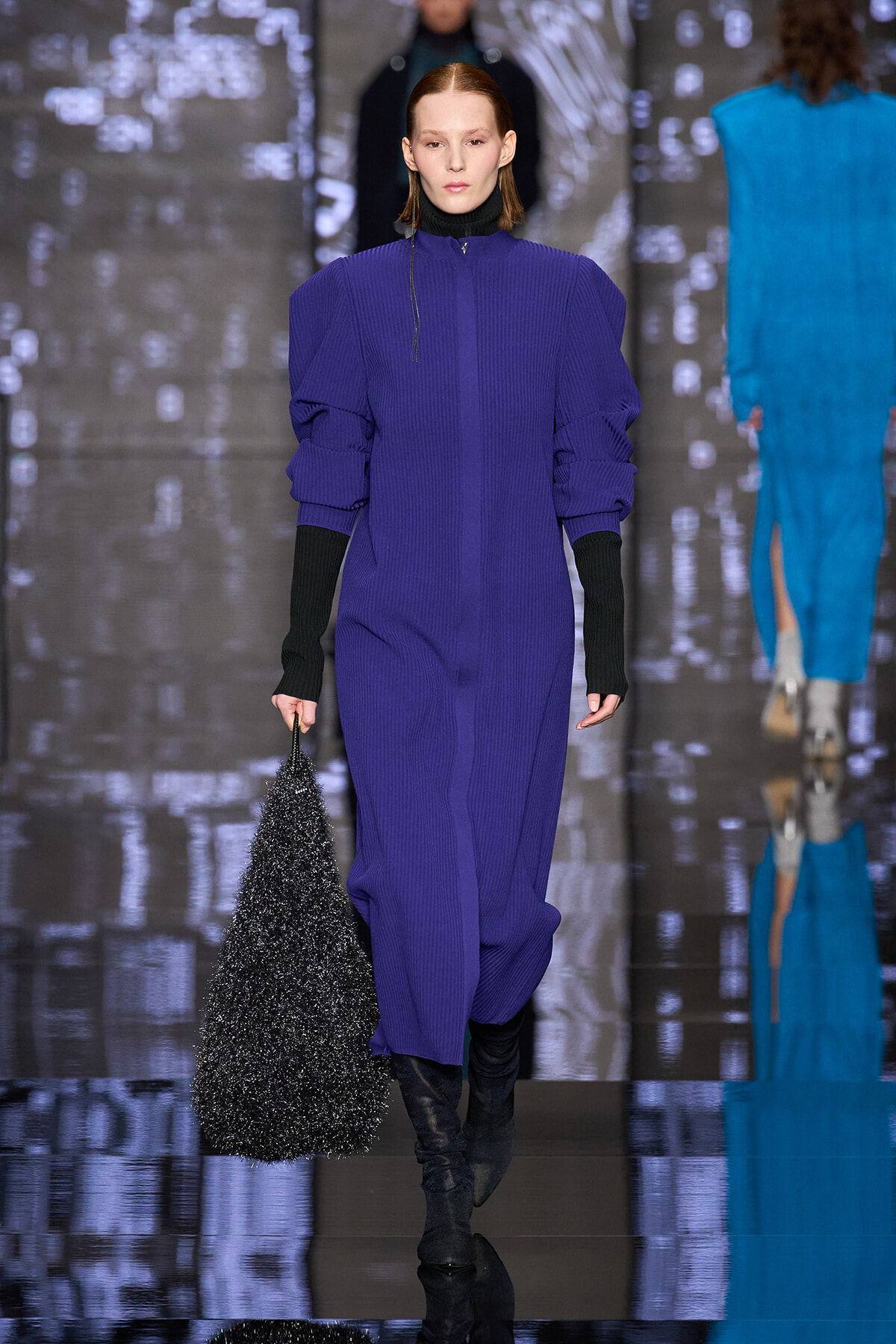 Model walking runway in a vivid purple knit midi dress with black turtleneck sleeves, holding a sparkly black bag.