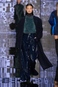 Model on a runway wearing a dark navy coat over a teal fluffy knit top and glossy navy wide-leg pants, with sleek straight hair.