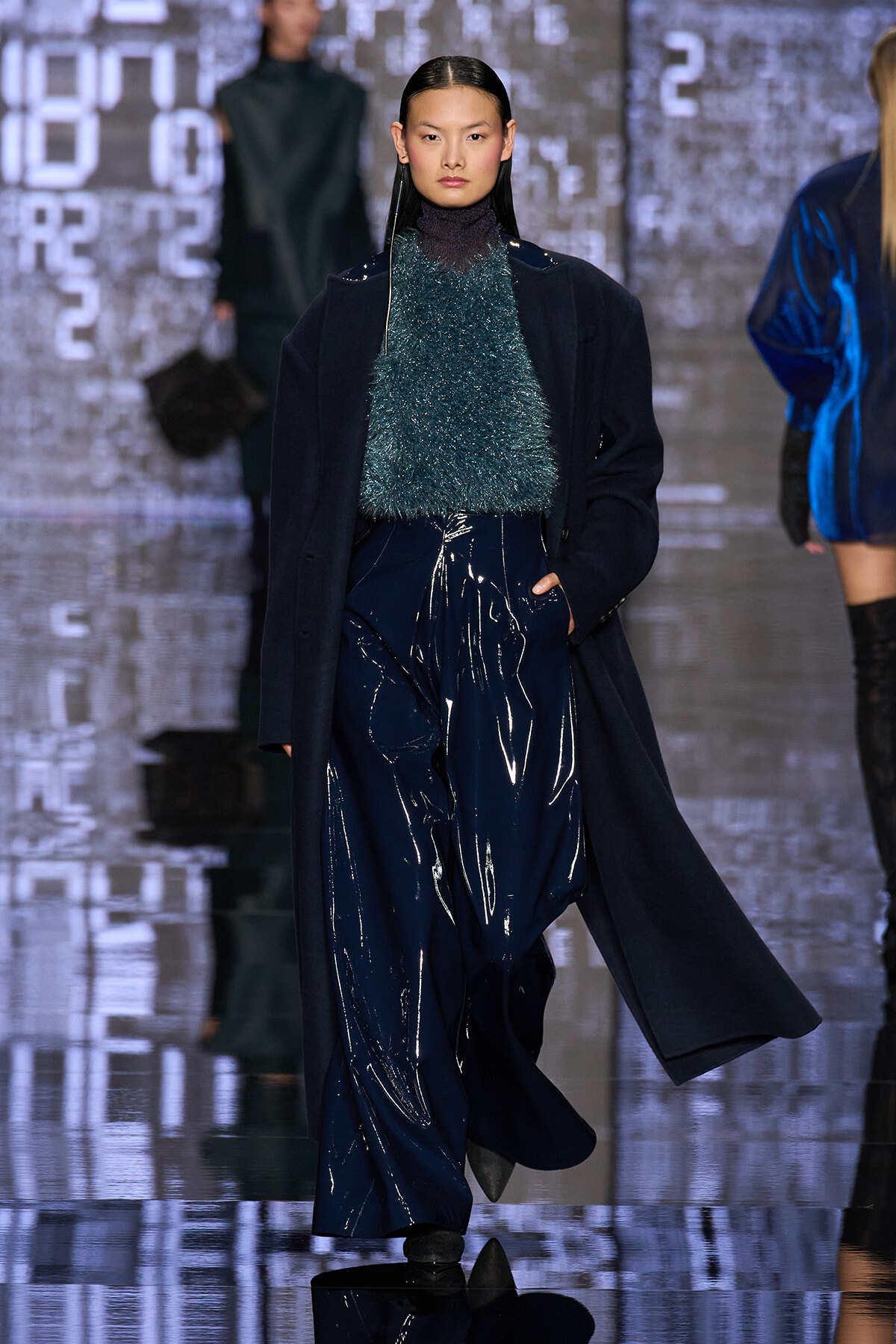 Model on a runway wearing a dark navy coat over a teal fluffy knit top and glossy navy wide-leg pants, with sleek straight hair.