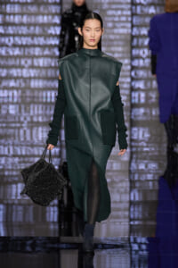Model walks the runway in a dark green oversized leather-look sleeveless coat with black knit sleeves, carrying a glittery black handbag.
