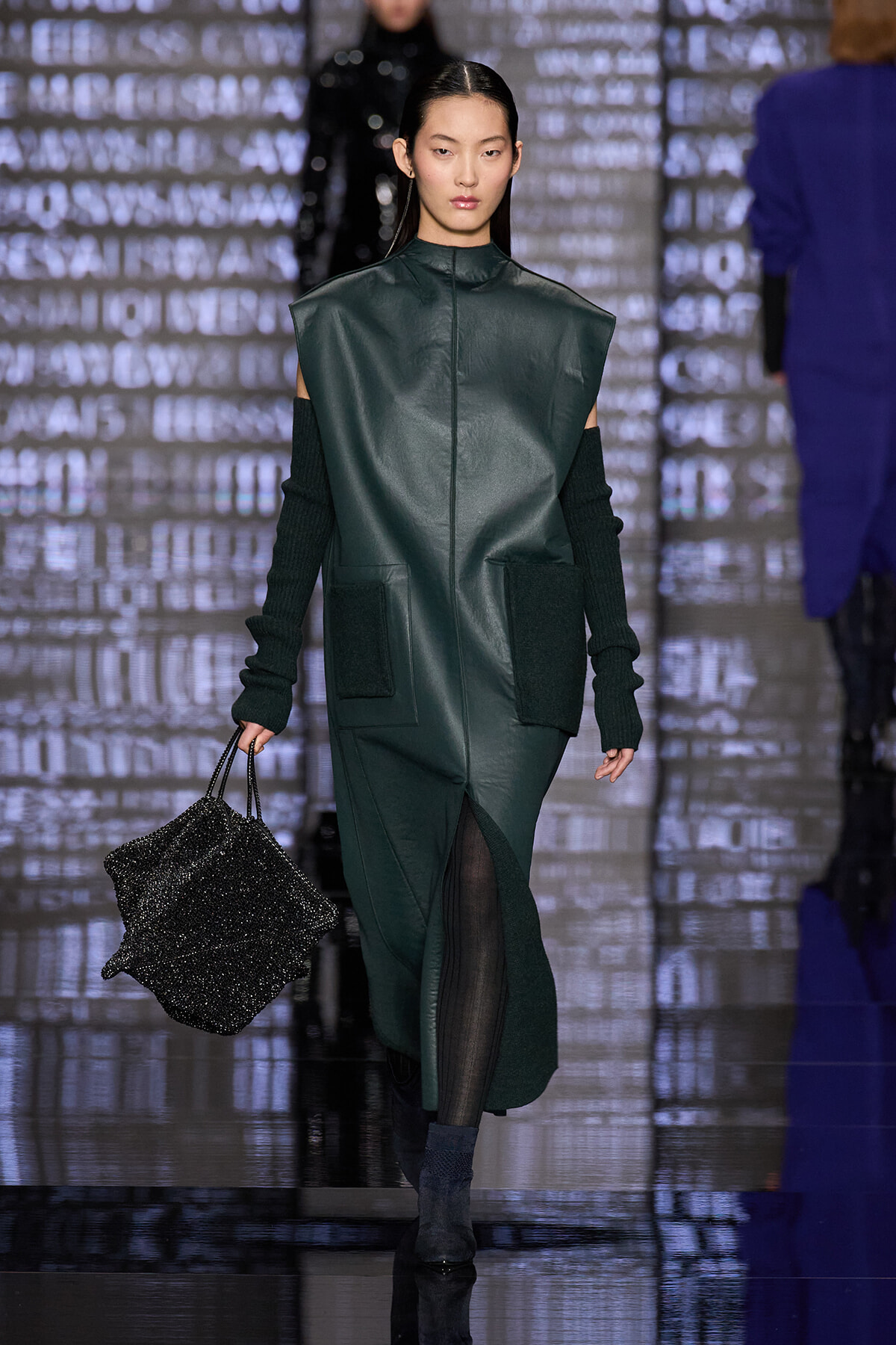 Model walks the runway in a dark green oversized leather-look sleeveless coat with black knit sleeves, carrying a glittery black handbag.