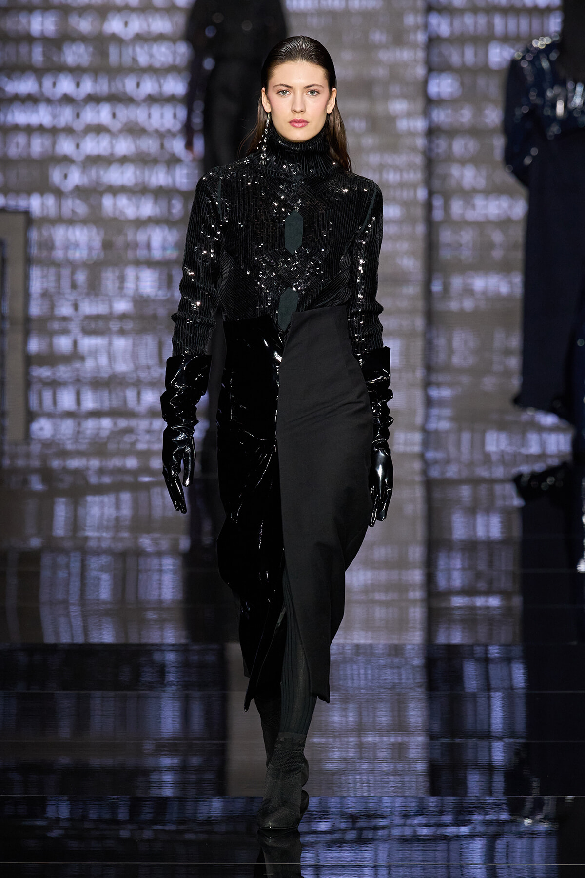 Model walking a fashion runway in a black sequined turtleneck and glossy black gloves with an asymmetric black skirt.