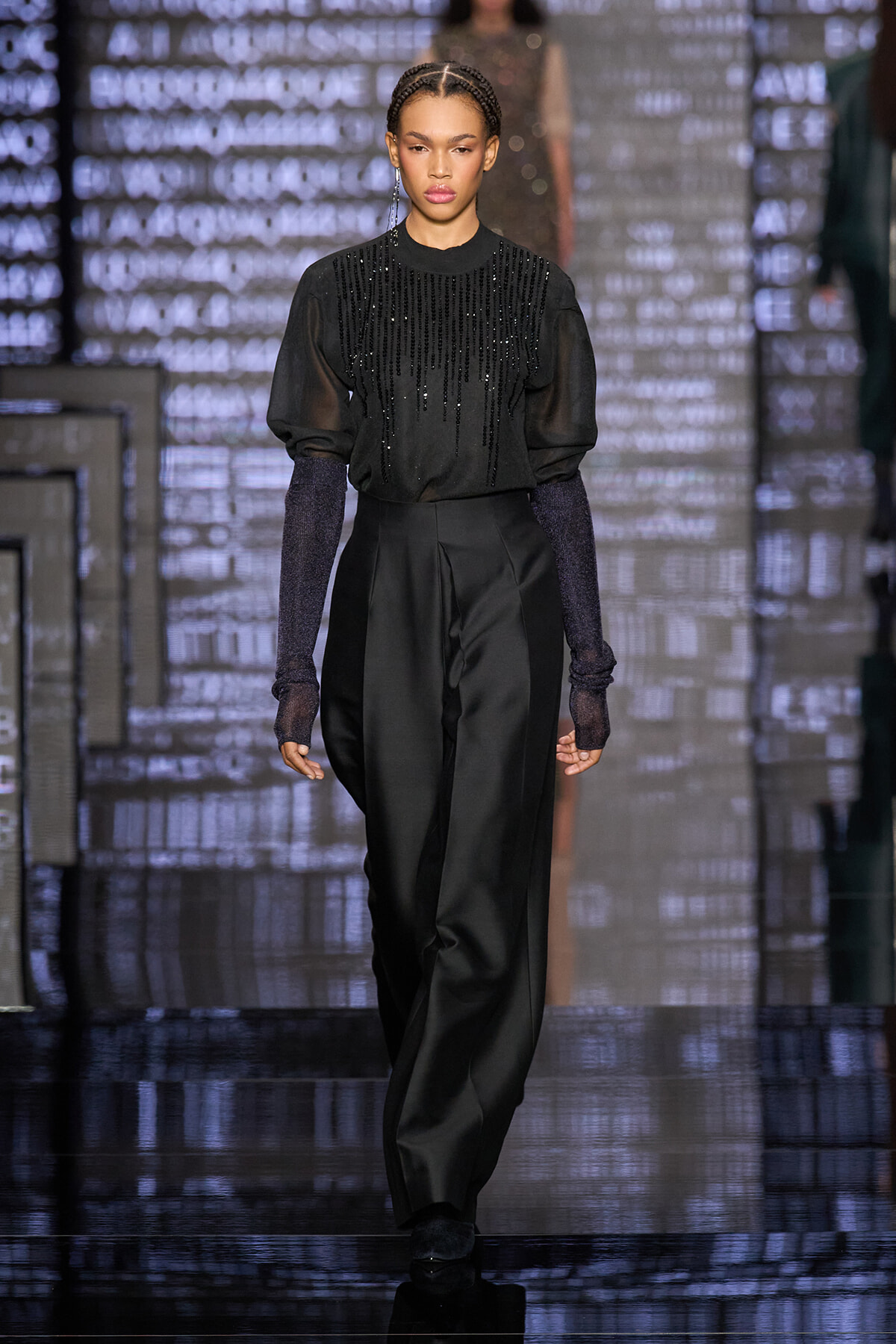 Model walks the runway in a black beaded top with sheer sleeves and tailored wide-leg pants.