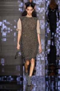 Model walks a glossy runway in a knee-length, glittery dark dress with textured metallic fabric and beige sheer sleeves; she carries a small beaded clutch.