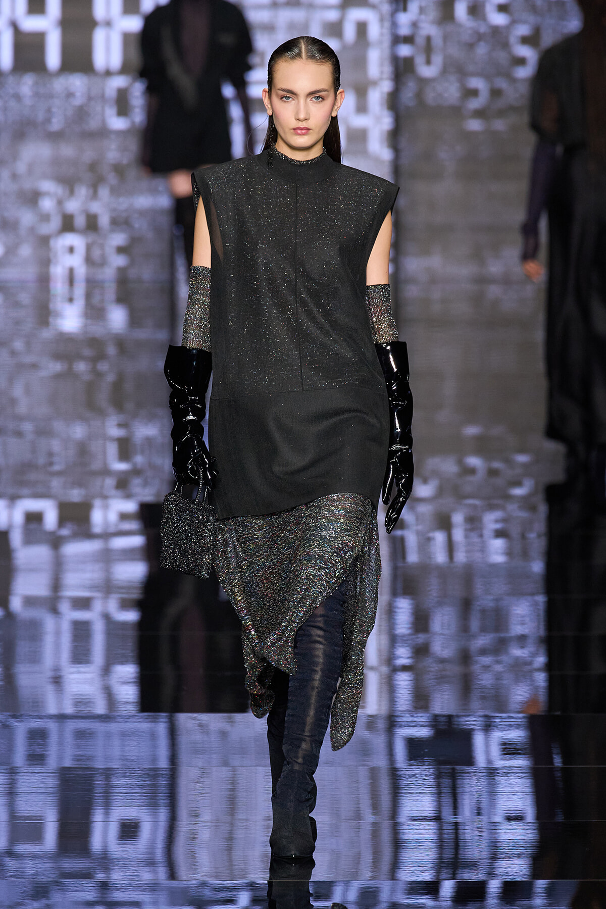 Model walks the runway in a glittery black ensemble with a sleeveless top, metallic skirt, and long shiny gloves.